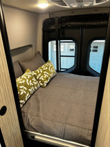 Cozy RV rental bedroom with a comfy bed, gray bedding, and green leaf-patterned pillows near small rear windows.