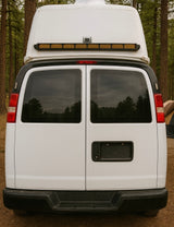 Rear view of a white rv rental van with a high roof, tinted windows reflecting trees, parked in a pine forest campsite.