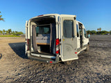 White Ford Transit camper van (RV rental) with open doors showing interior bed, parked on gravel in a sunny lot.