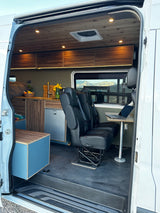 Modern RV rental camper van interior: wood ceilings, kitchen, bed, passenger seats with laptop table.