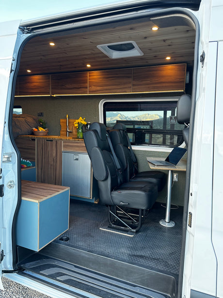Modern RV rental camper van interior: wood ceilings, kitchen, bed, passenger seats with laptop table.