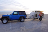 Blue Jeep Wrangler towing a silver travel trailer on concrete by a sandy beach and ocean. Great for an rv rental!