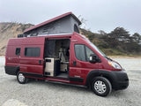 Red pop-top RV rental camper van with open side door revealing its interior, parked on gravel by a coastal hill.