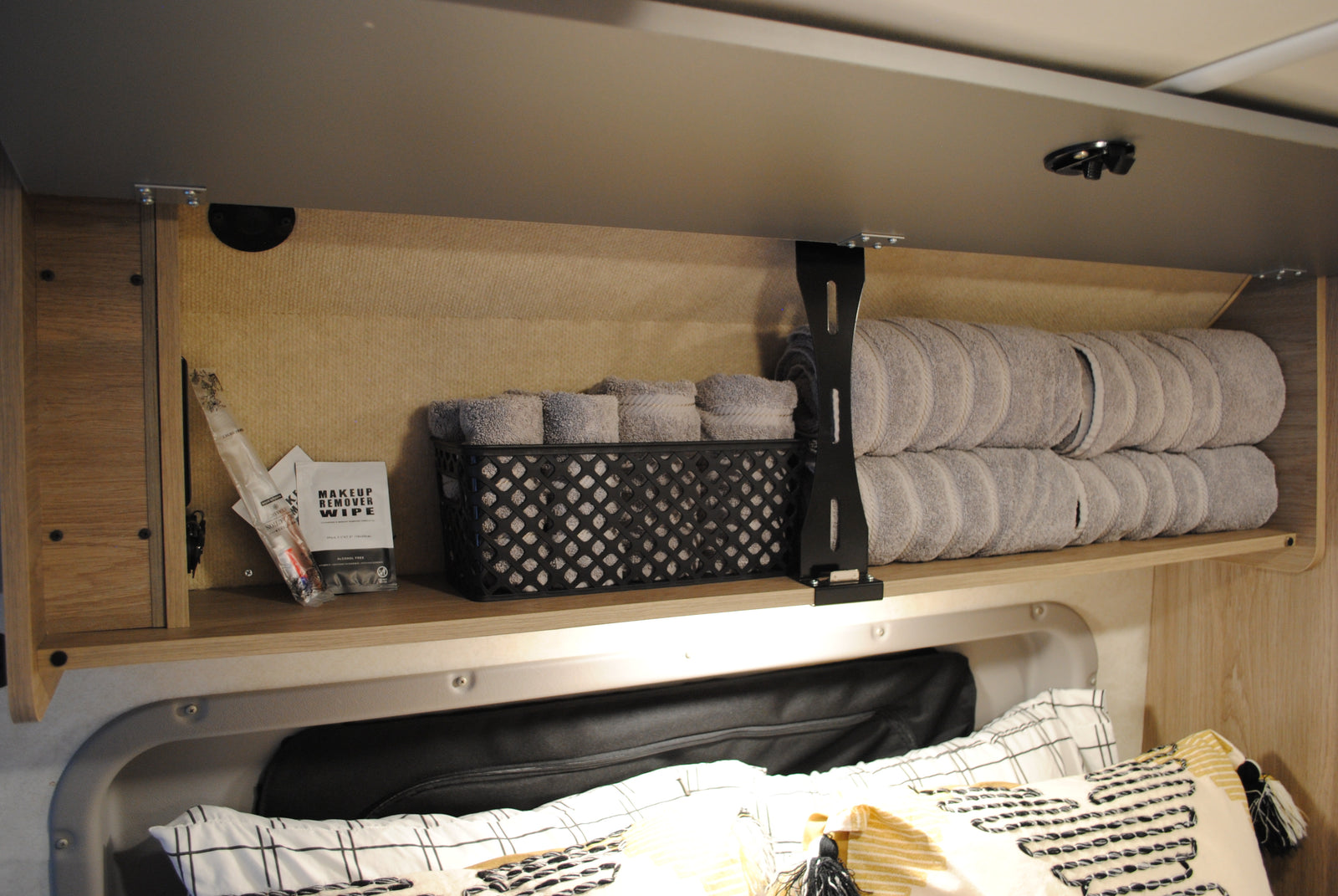 RV rental overhead storage: neatly folded grey towels, toiletries, and compact organization above comfortable bedding.