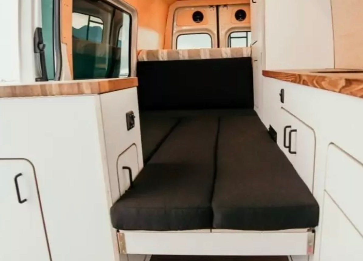 RV rental: Camper van interior shows a cozy black cushioned bed, white cabinets, and wood counters for comfy travel.