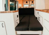 RV rental: Camper van interior shows a cozy black cushioned bed, white cabinets, and wood counters for comfy travel.
