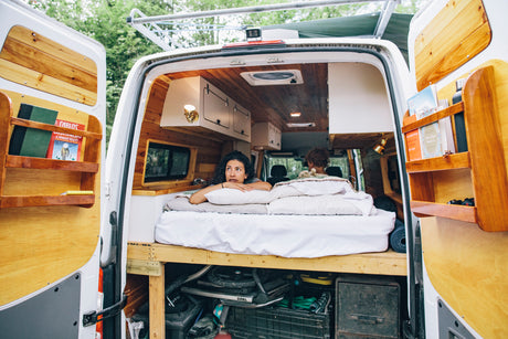 Inside a cozy RV rental: custom wood interior, bed with two people, and practical storage, ideal for van life.