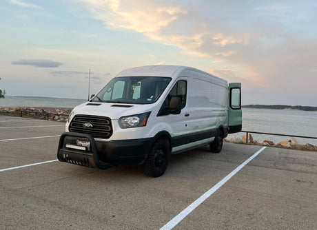 An RV rental ready for adventure: white Ford Transit camper van parked lakeside at dusk with the back door open, perfect for van life.