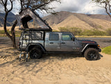 Experience outdoor adventure! This gray Jeep Gladiator, an ideal rv rental, has a roof-top tent & ladder, parked by mountains.