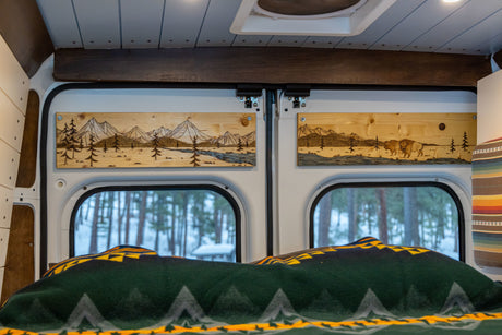 Cozy RV rental interior with custom wood art of mountains & bison, offering snowy forest views from the bed.