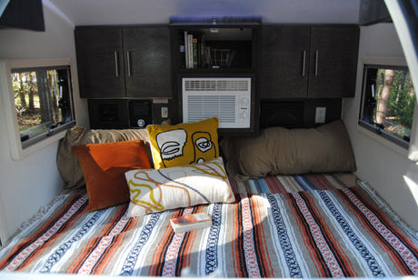 Inside a cozy RV rental: a comfortable bed with pillows, shelving, and an AC unit for travel.