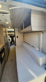 Modern RV rental interior with light wood cabinetry, a long grey fabric bench/bed, bright lighting, and vinyl flooring.