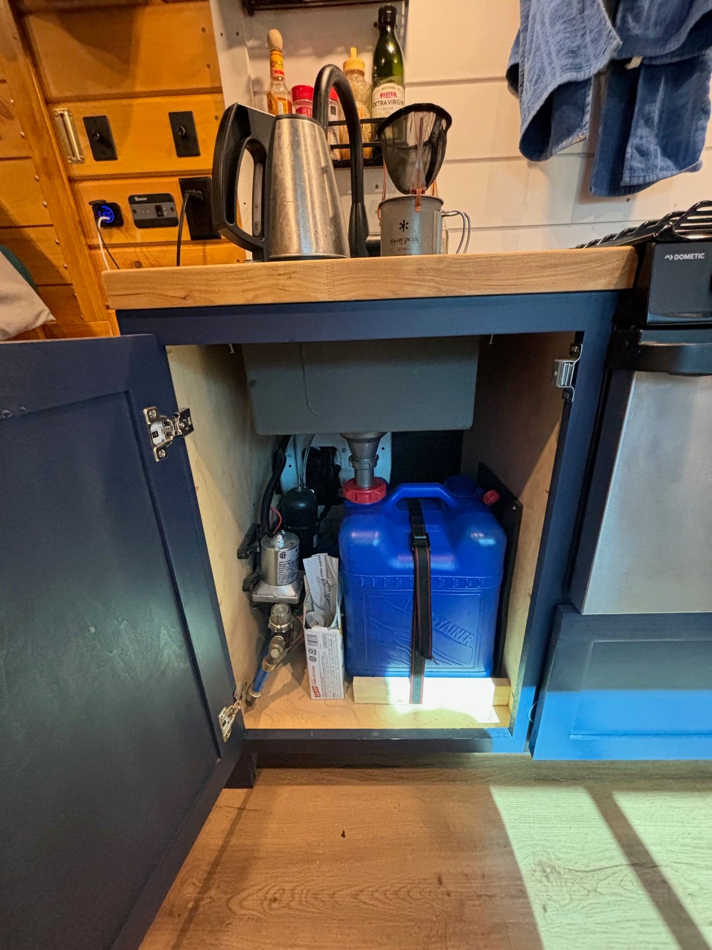 This RV rental galley features a kettle, stove, and an open cabinet revealing the compact water system with a pump & blue tank.