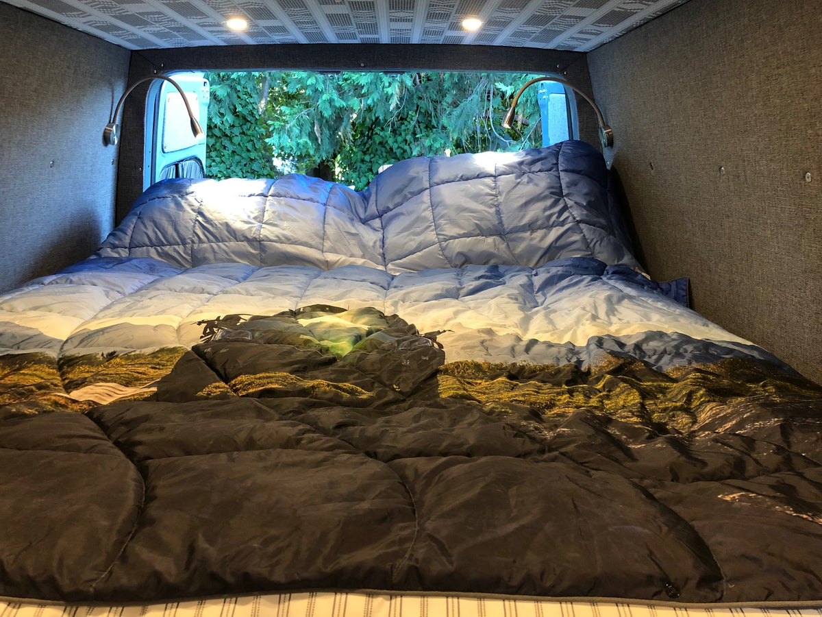 RV rental interior: cozy bed with mountain-themed comforter, reading lights, and view of lush trees. — photo 13