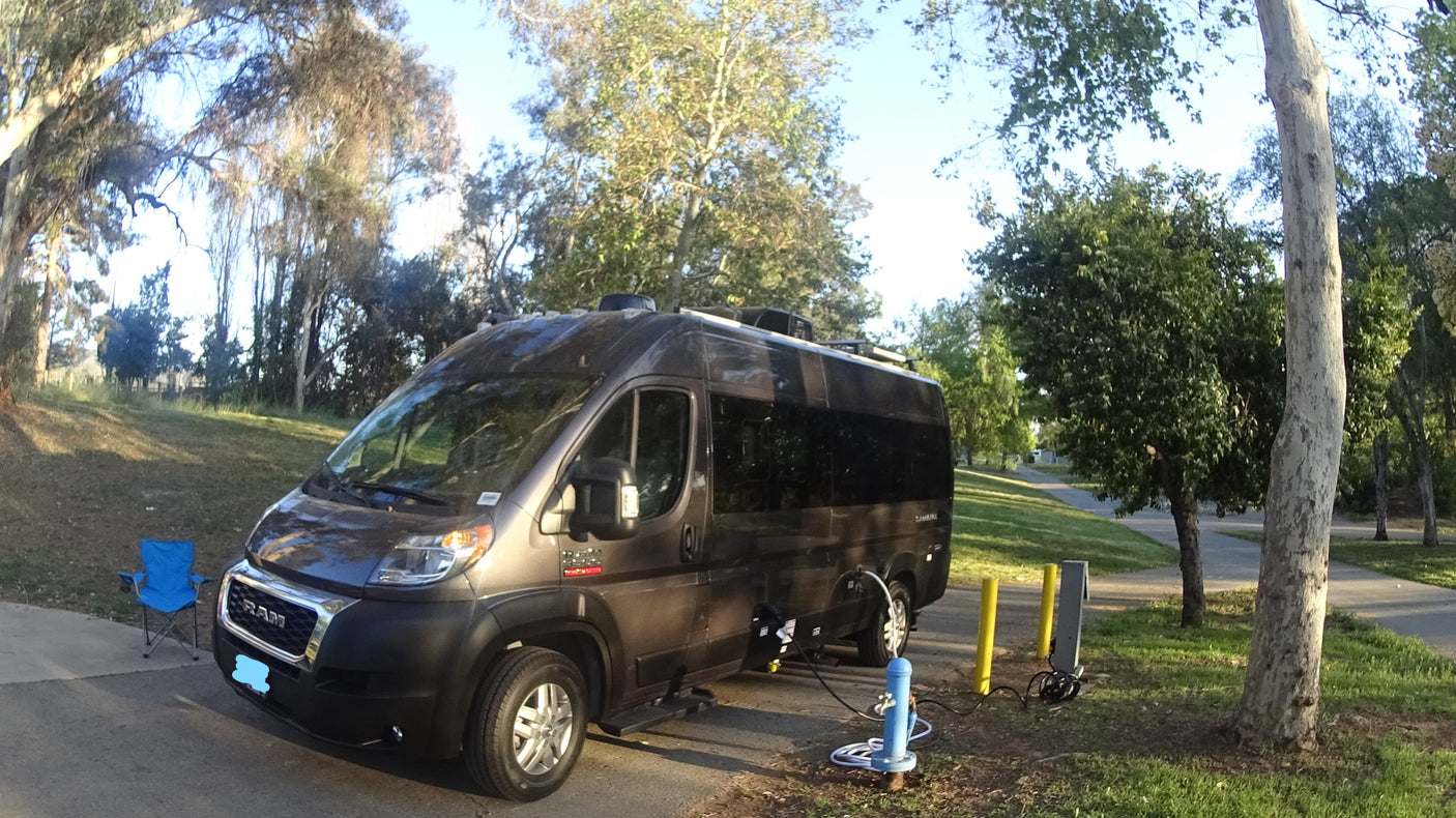 An RV rental, a dark grey campervan, is connected to campsite hookups on a sunny day. A blue chair waits beside it.