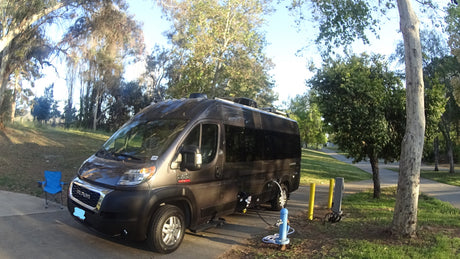 An RV rental, a dark grey campervan, is connected to campsite hookups on a sunny day. A blue chair waits beside it.