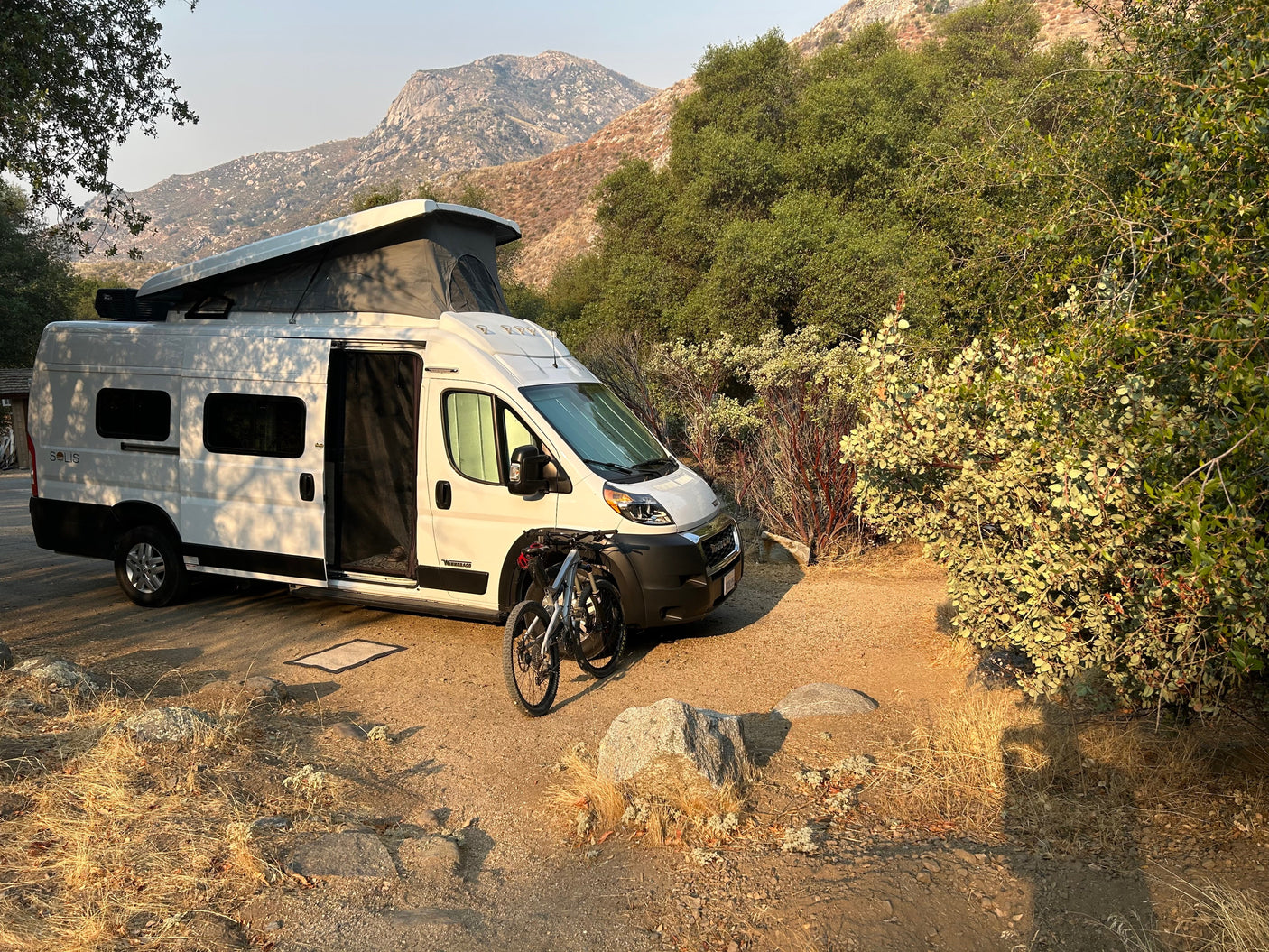 A white Winnebago camper van, an RV rental, with two mountain bikes parked at a scenic, rustic mountain campsite.