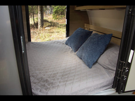 Inside this inviting RV rental: a comfortable bed with blue pillows and a large window overlooking a peaceful forest.
