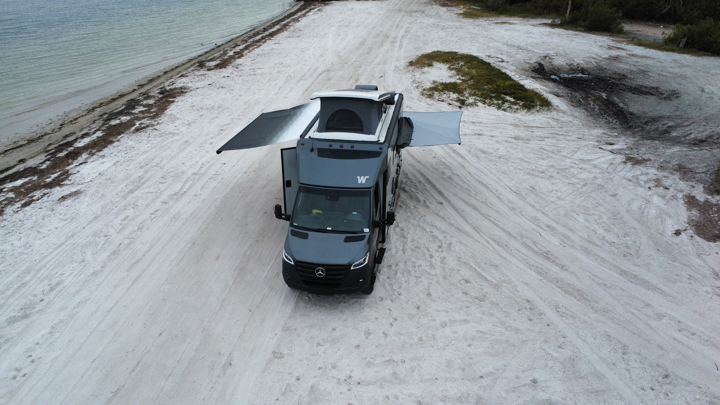 Grey RV rental van on a wide sandy beach. Its pop-top and side awnings are extended for a comfortable, scenic beach camp.
