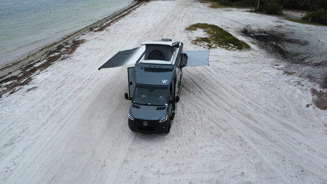 Grey RV rental van on a wide sandy beach. Its pop-top and side awnings are extended for a comfortable, scenic beach camp.