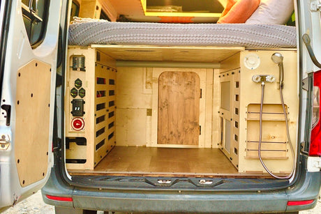RV rental camper van's organized rear. Features modular plywood storage, electrical panel, and shower unit below a comfy bed.