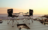 Two black folding chairs on a deck, overlooking a sunset ocean with distant surfers. Perfect RV rental view for a coastal escape.