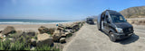 Coastal RV rental parked with an open door next to a sandy beach. Waves, other RVs, and hills complete the sunny scene.