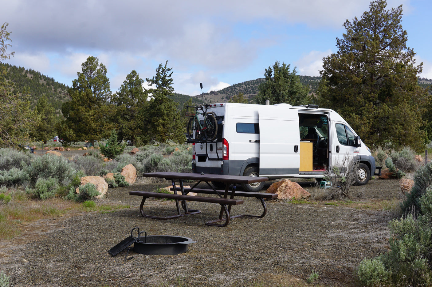 An rv rental van is parked at a scenic campsite with a picnic table and fire pit, surrounded by trees and mountains.