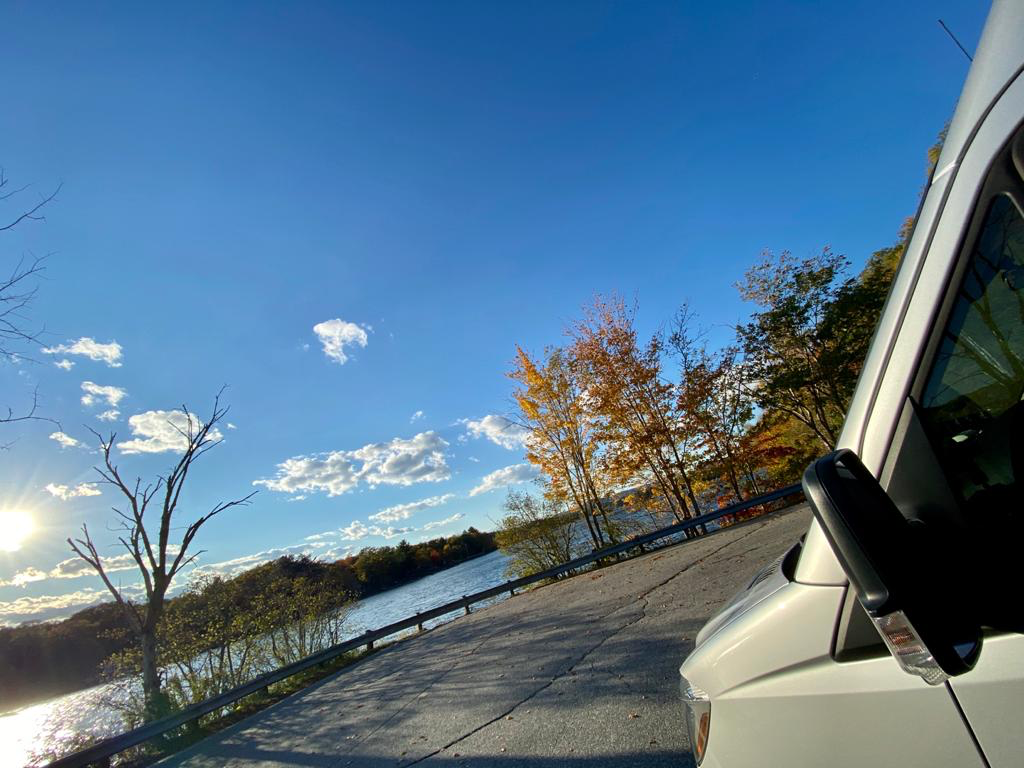 A white RV rental on a road beside a sunlit river and colorful fall trees. Blue sky with clouds, highlighting a peaceful autumn travel scene.