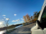 A white RV rental on a road beside a sunlit river and colorful fall trees. Blue sky with clouds, highlighting a peaceful autumn travel scene.