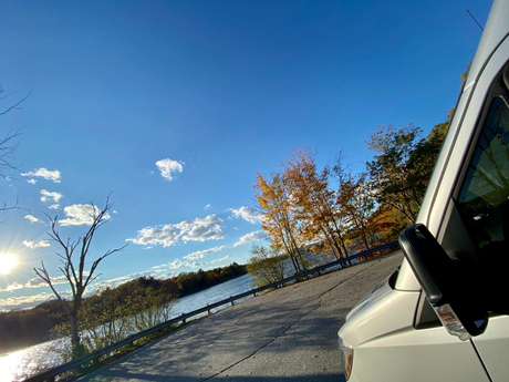 A white RV rental on a road beside a sunlit river and colorful fall trees. Blue sky with clouds, highlighting a peaceful autumn travel scene.