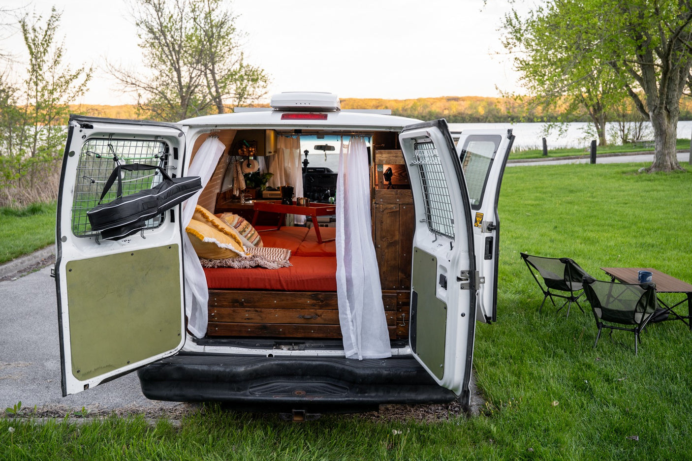 An RV rental's back doors are open, revealing a cozy van life interior with a bed, ready for a serene outdoor trip.