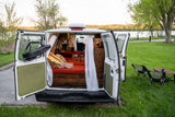 An RV rental's back doors are open, revealing a cozy van life interior with a bed, ready for a serene outdoor trip.