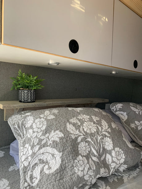 Cozy RV rental bedroom showcasing smart overhead storage, a small plant on a wood shelf, and soft floral bedding.