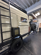 This custom light-colored RV rental van features a roof rack, rear spare tire, and side-mounted utility can, perfect for adventure.