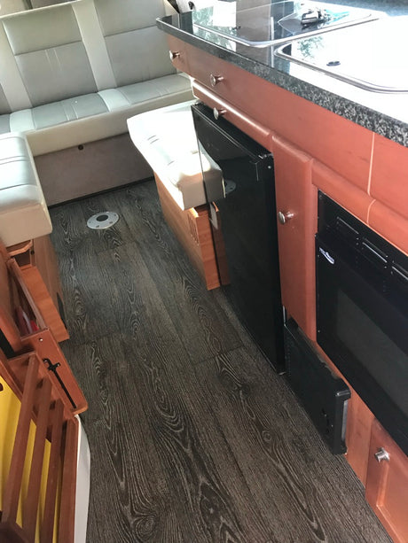 Compact rv rental interior with dark wood flooring, light seats, and a kitchenette featuring a sink, fridge, and microwave.