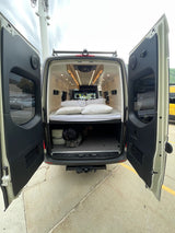 Open doors reveal a cozy bed & storage in this white camper van, ready for your RV rental adventure.