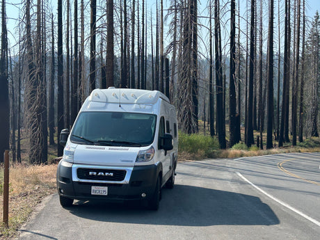 Scenic rv rental road trip stop: a white camper van parked by charred trees in a post-wildfire forest. California travel.