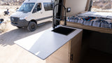 Inside an RV rental: a sleek kitchen counter with sink & faucet, and a comfortable bed. A Sprinter van is parked outside in nature.