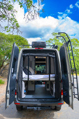 Open back doors of an RV rental showcase a cozy bed & gear storage, set against a vibrant outdoor backdrop for adventure.