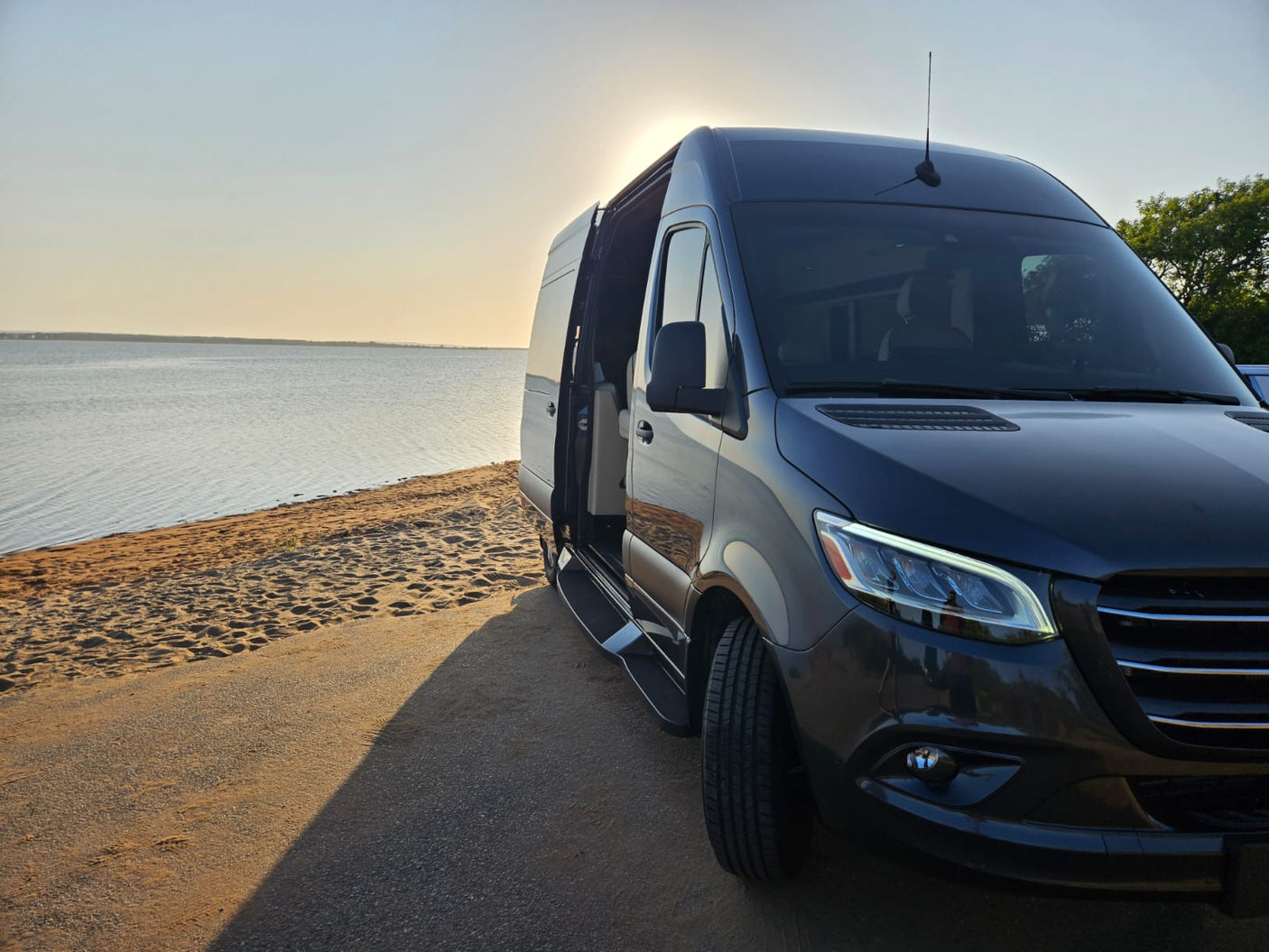 Dark camper van with open side door, parked on sandy ground by a lake at sunset. Ready for your RV rental adventure!