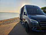 Dark camper van with open side door, parked on sandy ground by a lake at sunset. Ready for your RV rental adventure!