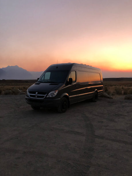 Dark Dodge Sprinter RV rental van silhouetted against a vibrant desert sunset with distant mountains.