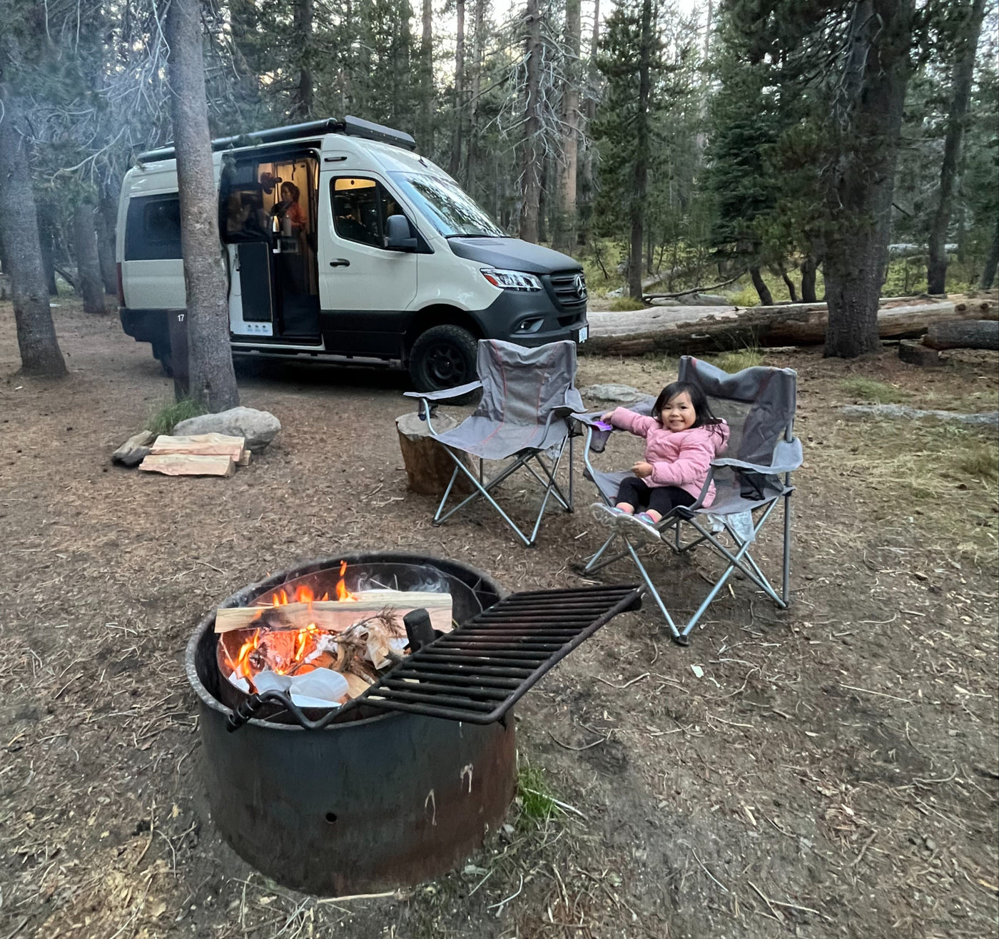 parked at a wooded campsite with an inflatable boat nearby.|RV rental camper van parked in a sunny forest campground. Tall trees surround the peaceful campsite.|Winnebago Revel RV rental set up at a sunny forest campsite with adventure gear on the ground.|A joyful child peeks from a light-colored RV rental window — photo 5