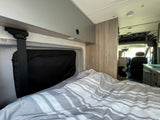 Cozy interior of an RV rental: a bed with striped bedding, overhead storage, and the front cabin of this camper van.