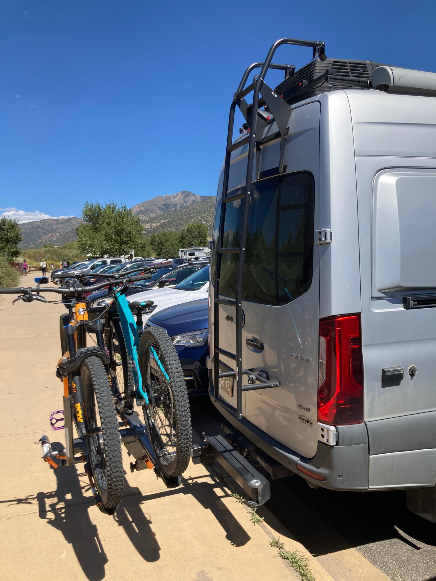 An RV rental van, a silver Mercedes Sprinter, sports two mountain bikes on its rear rack, amidst a sunny mountain backdrop.
