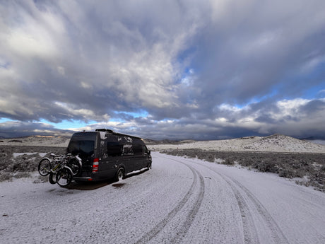 Dark gray RV rental with bikes on a snowy desert road. Dramatic winter clouds loom over snow-dusted hills.