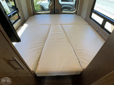 Inside an RV rental, showing a cream-colored convertible bed/seating area with multiple cushions and windows on both sides.