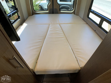 Inside an RV rental, showing a cream-colored convertible bed/seating area with multiple cushions and windows on both sides.