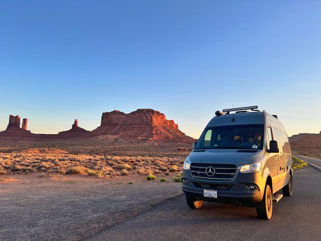 A gray Mercedes Sprinter RV rental van sits on a desert road, facing Monument Valley's red rock formations at sunset.
